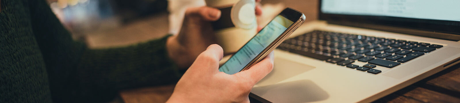 Woman using phone, holding coffee and in front of laptop at cafe.